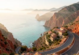 Route panoramique sur la côte ouest de la Corse, avec une mer turquoise, des falaises rouges et un village méditerranéen aux maisons en pierre.